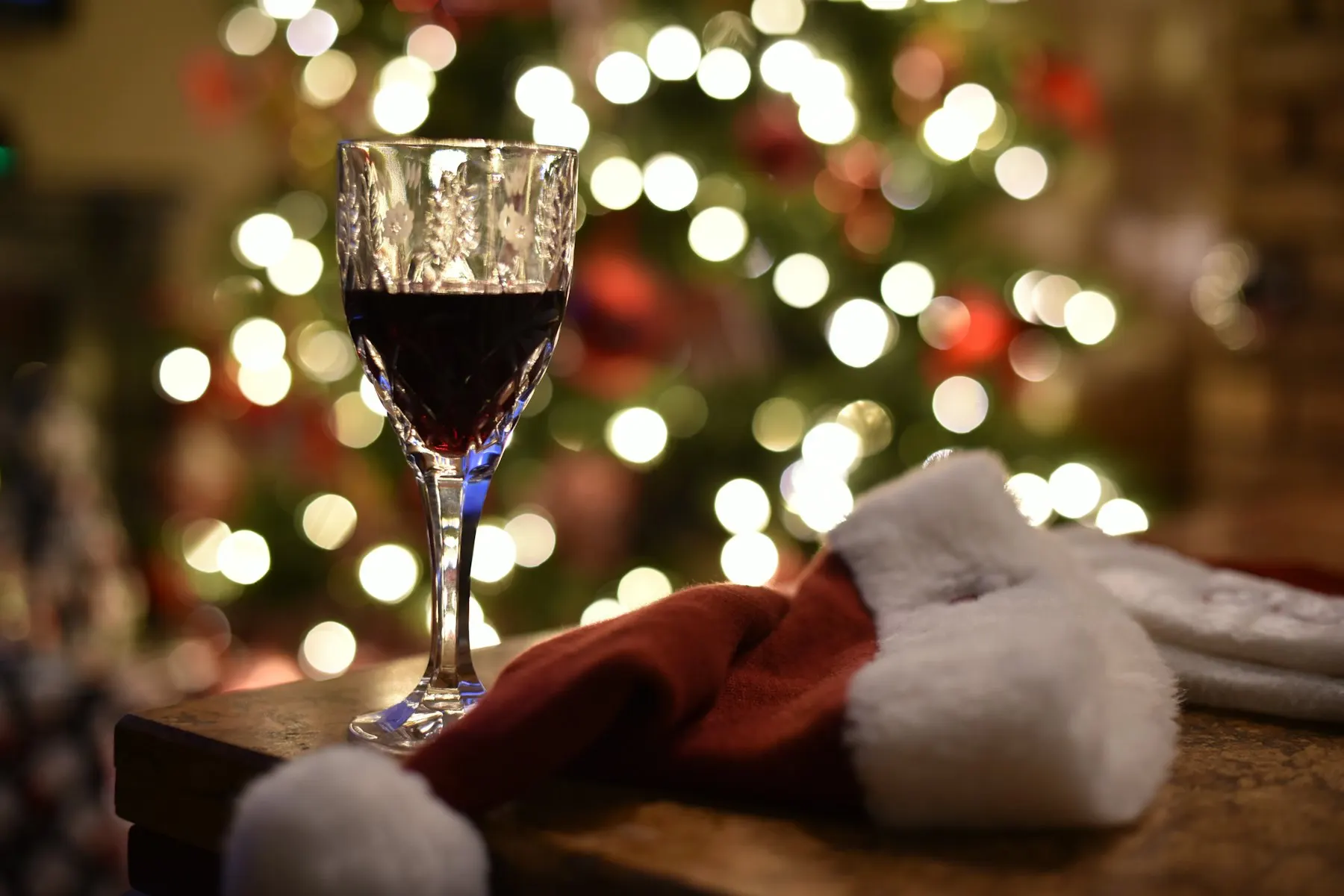 A glass of red wine beside a Santa hat in front of a lit Christmas tree